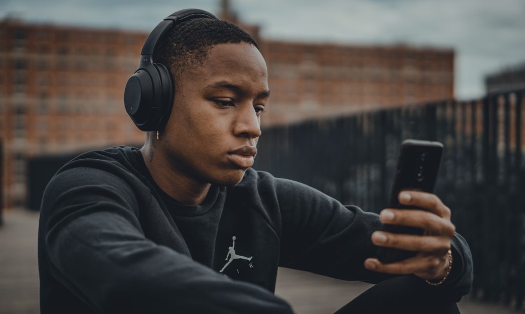 A man with headphones sits while holding his phone, searching for new music.